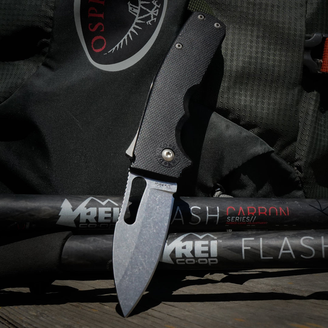 Shark-Cub slicer shark folding knife on a REI Flash Carbon Series hiking sticks with an osprey backpack in the background.