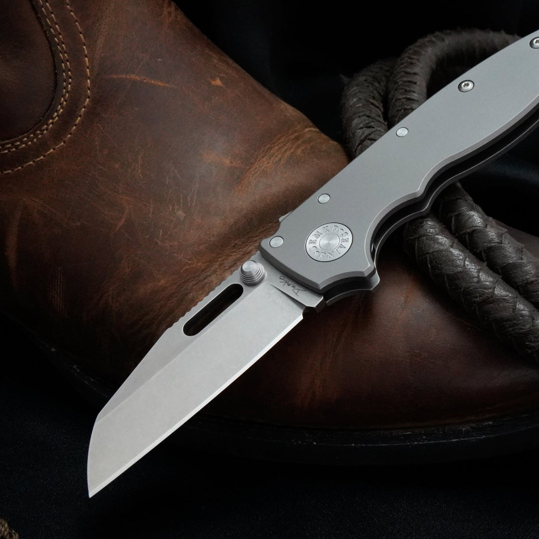 AD20.5 CPM 20CV shark foot folding knife with a smooth titanium handle on a boot with leather rope.