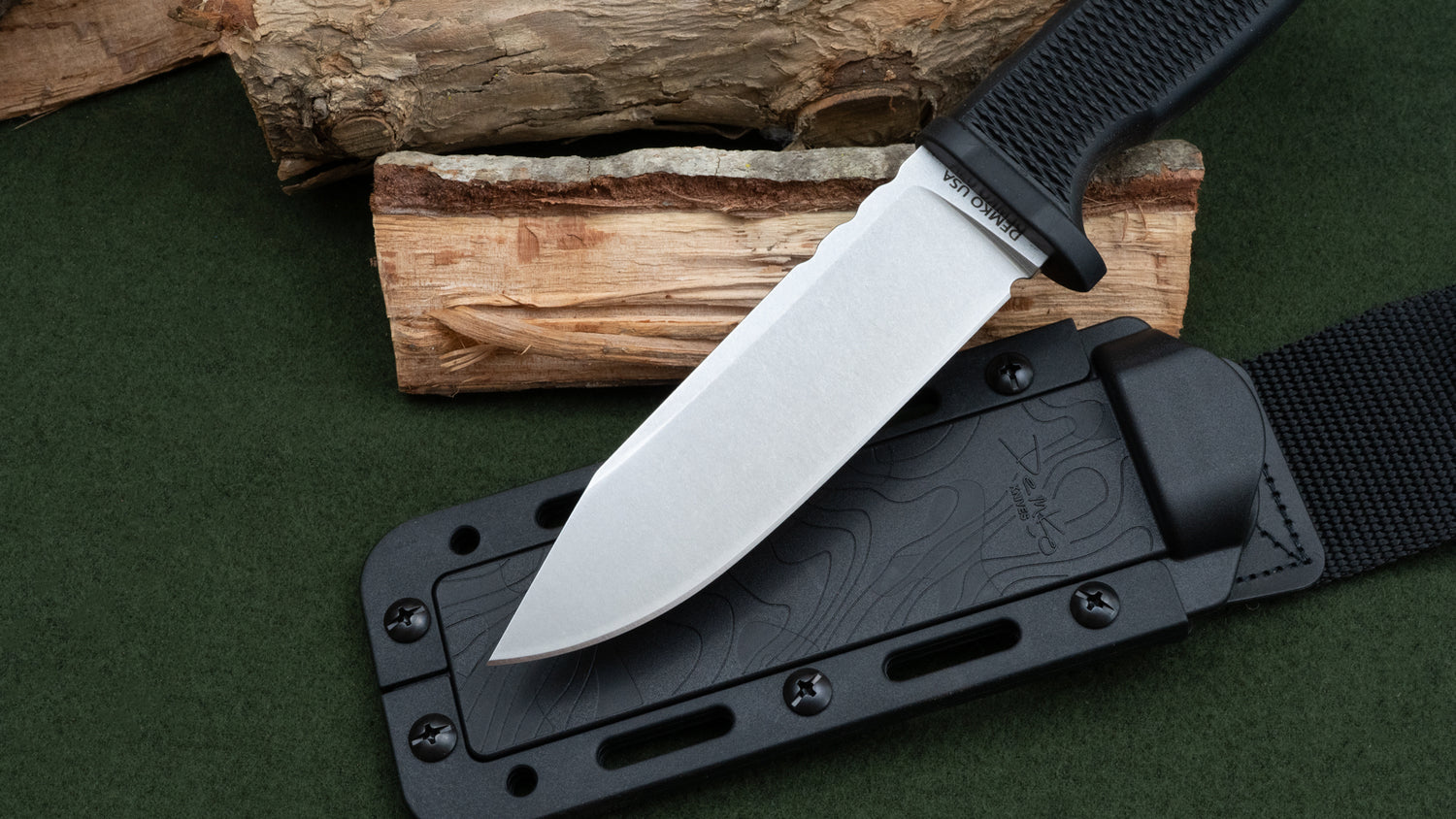 American made 3V clip point fixed blade knife with a black sheath on a green surface with wooden logs in the background.